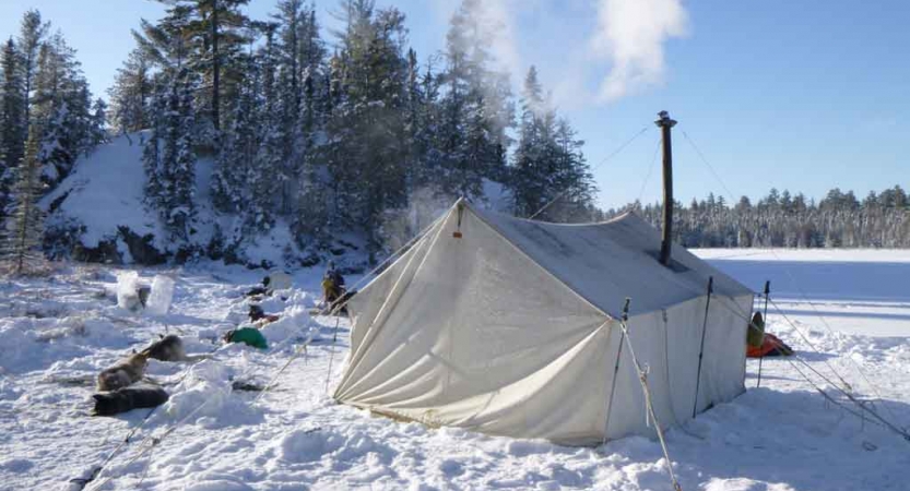 A tent with a small chimney emitting smoke sits on a frozen, snowy lake. Sled dogs rest in the snow nearby. 